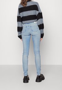 Donna che indossa jeans skinny blu chiaro, scarpe nere chunky e un maglione a righe grigio e nero, in piedi contro uno sfondo bianco semplice.