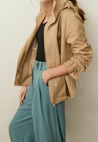 Woman wearing tan hooded jacket over black top and teal high-waist wide-leg pants with hand in pocket, standing against plain background.