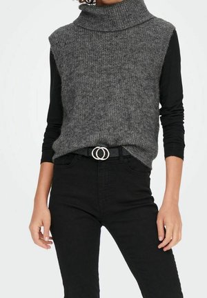 Personne portant un pull à col roulé gris sans manches par-dessus une chemise noire à manches longues, associé à un pantalon noir taille haute et une ceinture avec boucle à anneau double.