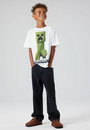 Witte t-shirt met een groene pixelachtige Creeper-karakter uit Minecraft, gecombineerd met donkere spijkerbroek en bruine instap schoenen.