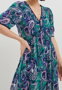 Sukienka w kwiatowy wzór paisley w kolorach zielonym, niebieskim i purpurowym. Dekolt w serek, krótkie rękawy, marszczona talia, lekki materiał o miękkiej teksturze.