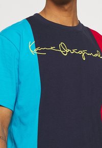 Tričko s krátkým rukávem s námořnickými, tyrkysovými a červenými panely; zlatě vyšívané logo "Kain Original" na hrudi; měkká bavlněná textura.