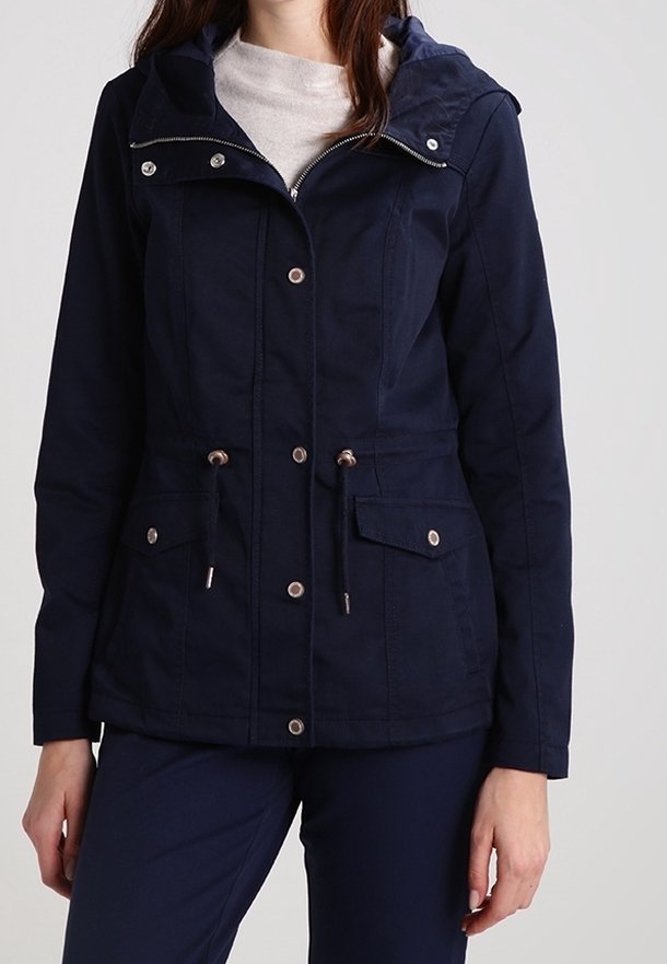 Femme portant une veste à capuche bleu marine avec fermeture éclair à l'avant, boutons-pression, taille coulissée avec cordon et poches à rabat sur un pantalon bleu marine assorti.