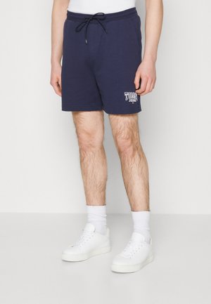 Marineblå bomuldsbukser med elastisk talje, justerbar snor og hvid logo-detalje. Sammen med hvide sneakers og sokker.