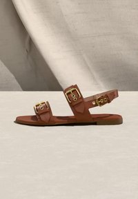 Sandale plate marron avec trois brides réglables à boucles dorées et une semelle lisse, présentée sur un fond neutre en tissu et surface.