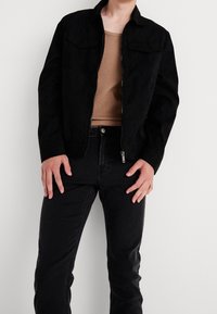 Musta mokkanahkajakki vetoketjulla, jossa on kaksi rintataskua. Yhdistetty ruskean topin ja mustien farkkujen kanssa.