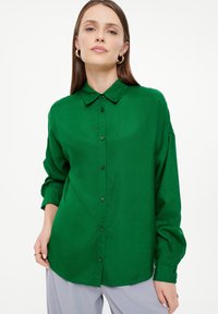 Grüne Bluse mit einem Knopfverschluss, weicher Textur, langen Ärmeln und einem klassischen Kragen. Verfügt über passende Knöpfe und einen abgerundeten Saum.