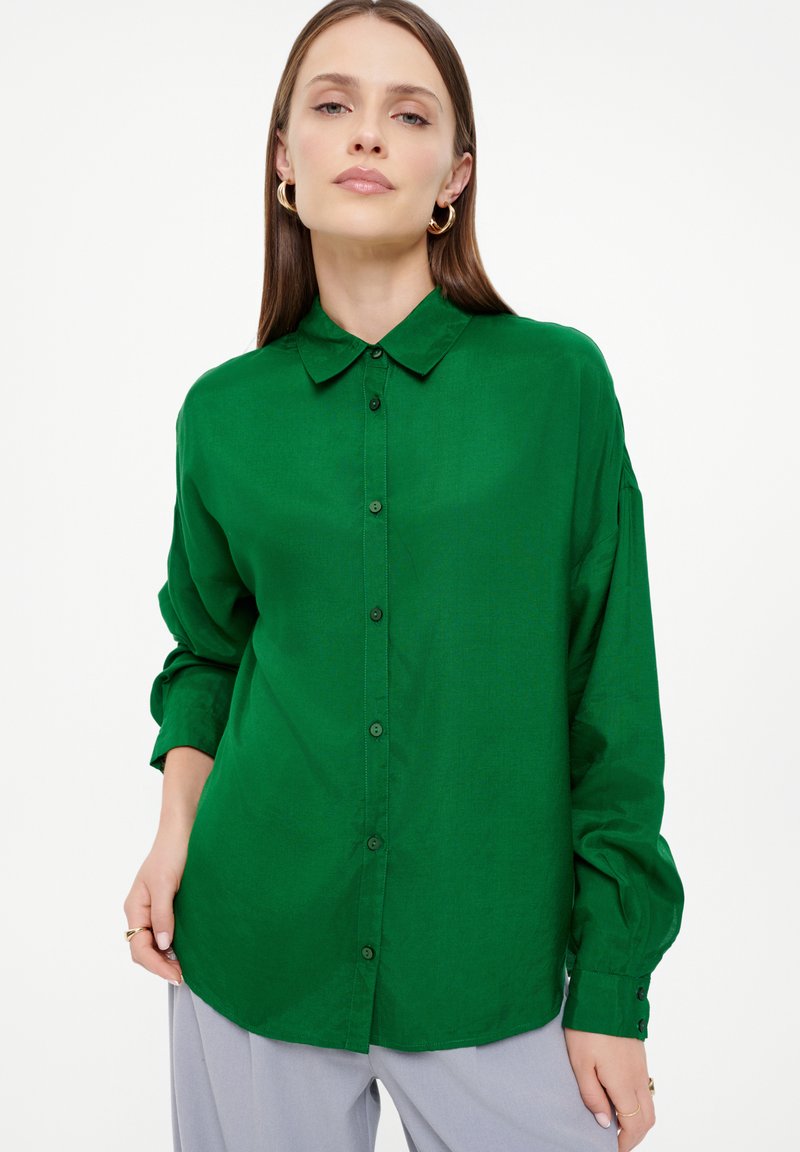 Grüne Bluse mit einem Knopfverschluss, weicher Textur, langen Ärmeln und einem klassischen Kragen. Verfügt über passende Knöpfe und einen abgerundeten Saum.