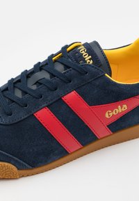 Zapatillas de ante azul marino con rayas rojas, interior amarillo y suela de goma. Presenta un logo dorado en el lateral y detalles amarillos en la lengüeta.