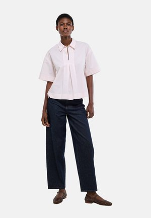 Une femme se tient debout, portant un chemisier ample à manches courtes de couleur rose pâle, un jean large sombre, et des chaussures Mary Jane en cuir tissé marron, devant un fond uni.