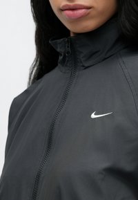 Μαύρο μπουφάν με φερμουάρ, κατασκευασμένο από ελαφρύ υλικό. Έχει ψηλό γιακά και μικρό λευκό λογότυπο Nike στο αριστερό μέρος.