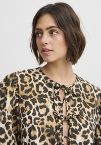 Blúzka s leopardím vzorom v béžovej a čiernej farbe, s okrúhlym výstrihom so stuhami a vypchávkami na rukávoch, vyrobená z ľahkej tkaniny.