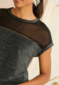 Top nero con parte superiore in mesh trasparente nera, caratterizzato da fili metallici e una sezione inferiore testurizzata. Campione di dettagli con pizzo delicato.