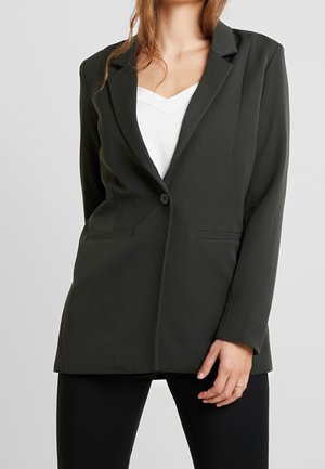 Femme portant un blazer noir ajusté sur un haut blanc, debout avec une main reposant près de la hanche sur un fond gris uni.