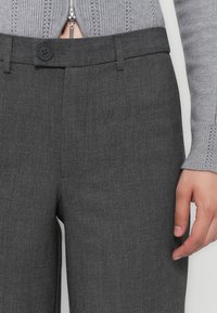 Pantaloni grigi su misura con una texture liscia, chiusura con un singolo bottone e passanti per cintura, abbinati a un maglione grigio lavorato a maglia.