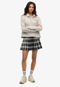 Grijze langwerpige polo met een witte kraag, gecombineerd met een zwart-wit geruite geplooide rok en beige sneakers met blauwe accenten.