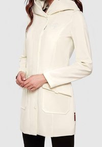 Kobieta w kremowym, wodoodpornym płaszczu przeciwdeszczowym z kapturem, przednimi kieszeniami i zatrzaskami, trzymająca ręce z przodu, na jednolitym tle.