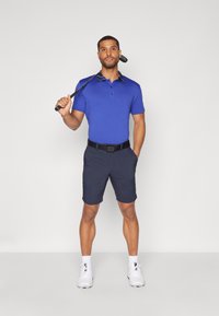 Modra polo majica z vzorčasto ovratnico, v kombinaciji s temno modrimi kratkimi hlačami. Bele športne čevlje dopolnjujejo obleko, ki jo drži golf palica.