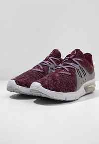 Chaussures de sport bordeaux et grises avec une tige tricotée, dotées de fermetures à lacets et d'une unité de coussin d'air visible dans la semelle. Semelle extérieure texturée.