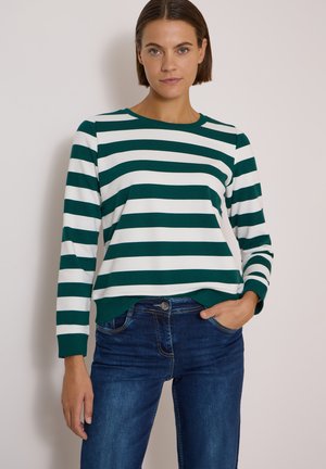 Femme aux cheveux courts et bruns portant un t-shirt à manches longues rayé horizontalement vert et blanc et un jean bleu, debout avec une main dans la poche.