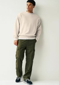 Sweatshirt beige clair avec col rond, coupe lâche ; pantalon cargo olive avec poches latérales ; baskets noires. Tenue simple et décontractée.