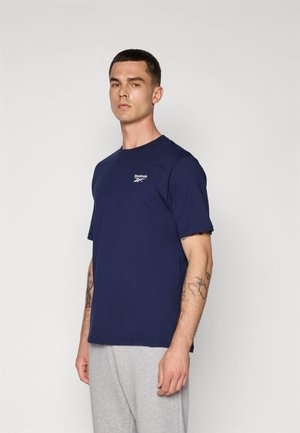 Mørkeblå bomulds T-shirt med korte ærmer og rund halsudskæring. Har et lille hvidt Reebok-logo på brystet. Bæres med grå svedbukser.