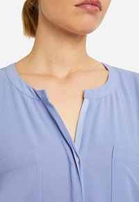 Primer plano del cuello y parte superior del pecho de una persona que lleva una blusa azul claro con escote redondo y detalle de botones en la parte delantera.