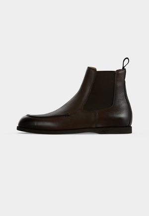 Dunkelbrauner Leder-Chelsea-Stiefel mit elastischen Seitenpaneelen, einer Zuglasche hinten und einer flachen schwarzen Sohle, im Profil vor weißem Hintergrund dargestellt.