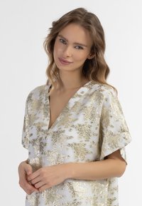 Bijela bluza s složenim zlatnim cvjetnim uzorkom, V-izrez, kratki rukavi i mekana, teksturirana tkanina. Model nježno spaja ruke.