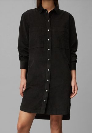 Kobieta ubrana w czarną sukienkę z długim rękawem na guziki, z dwoma kieszeniami na piersi, stojąca na tle jednolitego szarego tła.