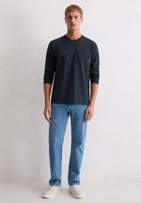 Granatowa koszulka z długim rękawem i kieszenią na piersi, zestawiona z jasnoniebieskimi dżinsami i beżowymi trampkami. Krój jest luźny i casualowy.