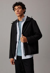 Giacca leggera nera con cappuccio, caratterizzata da una texture liscia. Sovrapposta a una camicia celeste, mostrata in un ambiente interno.