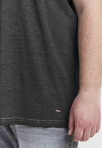 Tricou din bumbac gri închis cu o margine rotunjită, având un mic etichetă cu logo din cupru în partea stângă jos. Taietură casual, material texturat.