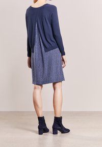 Top en tricot navy avec un pli dans le dos associé à une robe à motifs comprenant des carrés bleus. Le modèle porte des bottines navy, vue de dos.