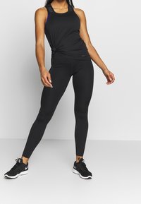 Top sportivo nero con dettaglio annodato e logo, abbinato a leggings neri aderenti e sneakers nere con dettagli bianchi.