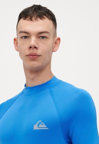 Mėlynas rash guardas, pagamintas iš elastingos medžiagos, turi aukštą apykaklę, plokščias siūles ir sidabrinį logotipą krūtinėje.