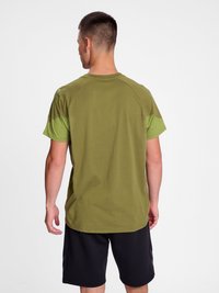 Homme de dos portant un t-shirt vert olive avec des accents vert clair sur les manches et un short noir sur un fond blanc uni.