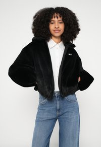 Γυναίκα με σγουρά μαλλιά που φοράει μαύρο χνουδωτό μπουφάν, λευκό πουκάμισο με κουμπιά και μπλε τζιν, στέκεται με τα χέρια στις τσέπες του μπουφάν.