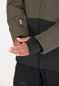 Olivgrüne und schwarze Jacke mit Frontreißverschluss. Verfügt über Klettverschlussmanschetten und ein weiches Innenfutter. Die Hände sind nah am Brustbereich zusammengelegt.