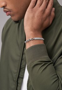 Nahaufnahme einer Person, die eine olivgrüne Jacke und ein silbernes Kettenarmband mit einer strukturierten rechteckigen Platte am Handgelenk trägt.