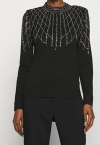 Pull noir à manches longues avec un col rond, orné de perles argentées formant un motif géométrique sur le buste et les épaules.