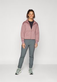 Light pink zip-up jacket with a stand collar, paired with dark gray tapered pants and green sneakers. Smooth fabric with subtle accents.