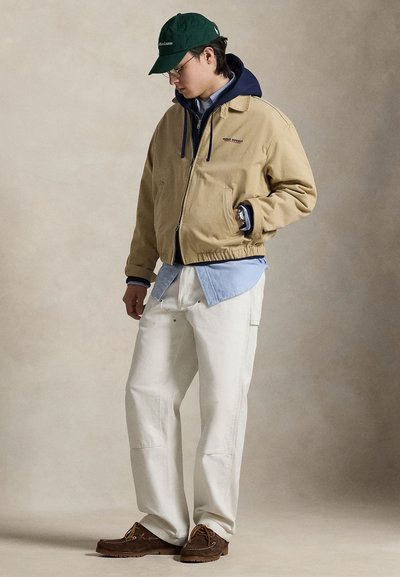 Μπεζ μπουφάν με επένδυση και μπλε σκουρόχρωμη κουκούλα, φορεμένο πάνω από ανοιχτό γαλάζιο πουκάμισο. Συνδυασμένο με εκρού παντελόνι και καφέ παπούτσια. Σκούφος σκούρου πράσινου χρώματος.