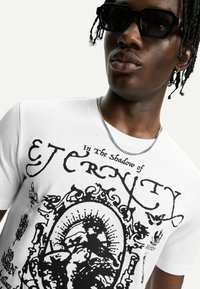 T-shirt in cotone bianco con stampa grafica nera che presenta testo e disegni intricati. Il modello indossa occhiali da sole neri e una catena d'argento.
