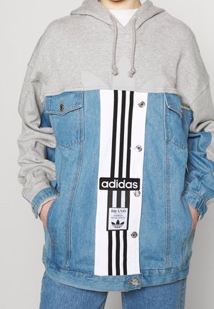Persoon in een blauwe spijkerjas met een grijze hoodie, aan de voorkant een wit paneel met zwarte Adidas-strepen en logo's, handen in de zakken.