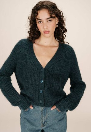 Gilet vert foncé texturé avec un décolleté en V profond, des manches longues et cinq grands boutons bleus sur le devant. Porté avec un jean taille haute.