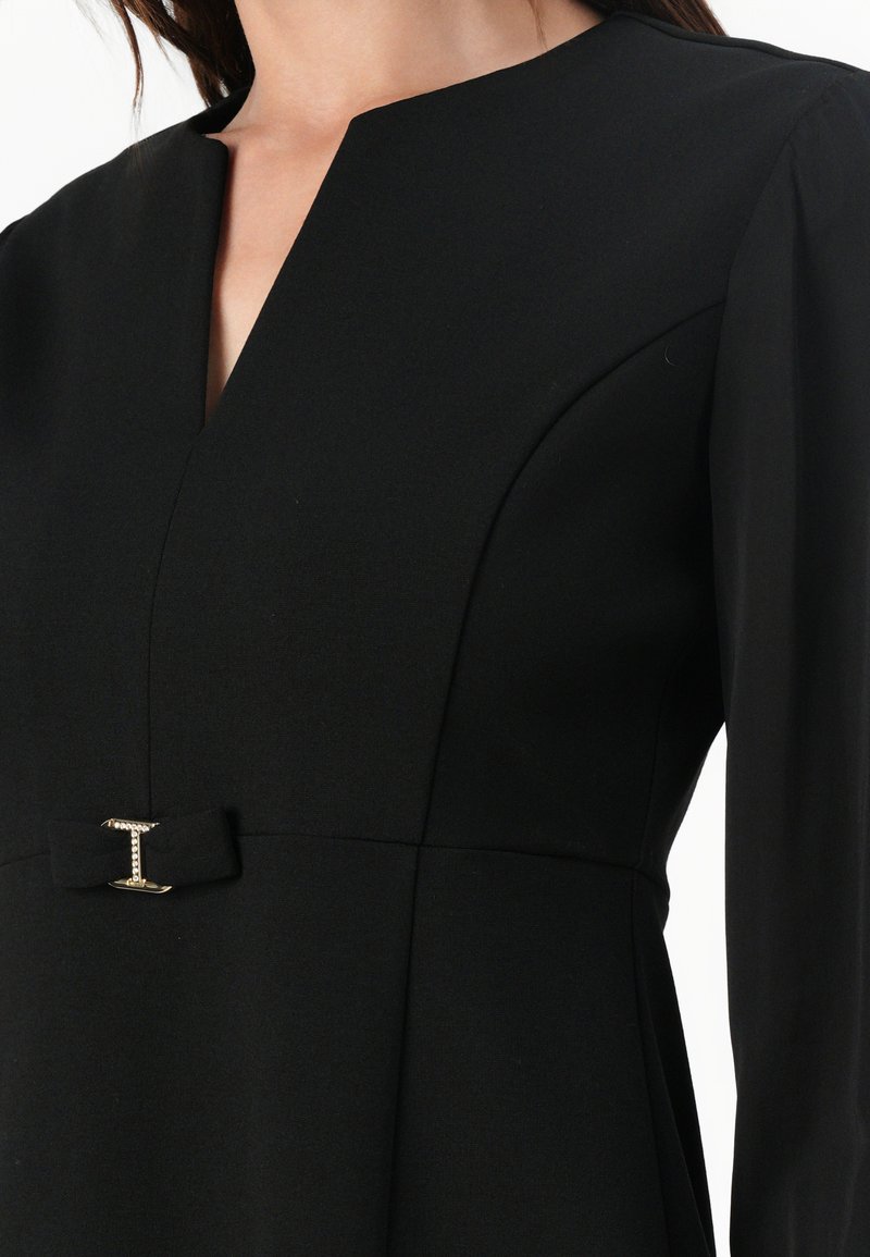 Abito nero con scollatura a V, maniche lunghe e corpino aderente. Dettaglio notevole include un fiocco con una chiusura metallica in vita.
