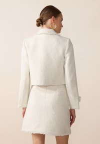 Giacca in tweed bianco con texture, dal design accorciato, dotata di colletto e maniche lunghe, abbinata a una mini gonna coordinata.
