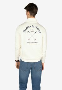Felpa bianca con grafica sul retro che mostra il testo "HARPER & NEYER", mazze da golf incrociate e la frase "EAST COAST OPEN." Indossata con jeans blu.