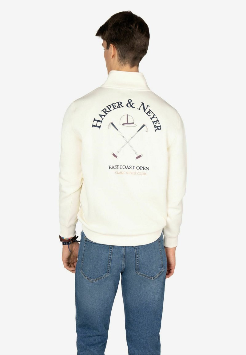 Felpa bianca con grafica sul retro che mostra il testo "HARPER & NEYER", mazze da golf incrociate e la frase "EAST COAST OPEN." Indossata con jeans blu.